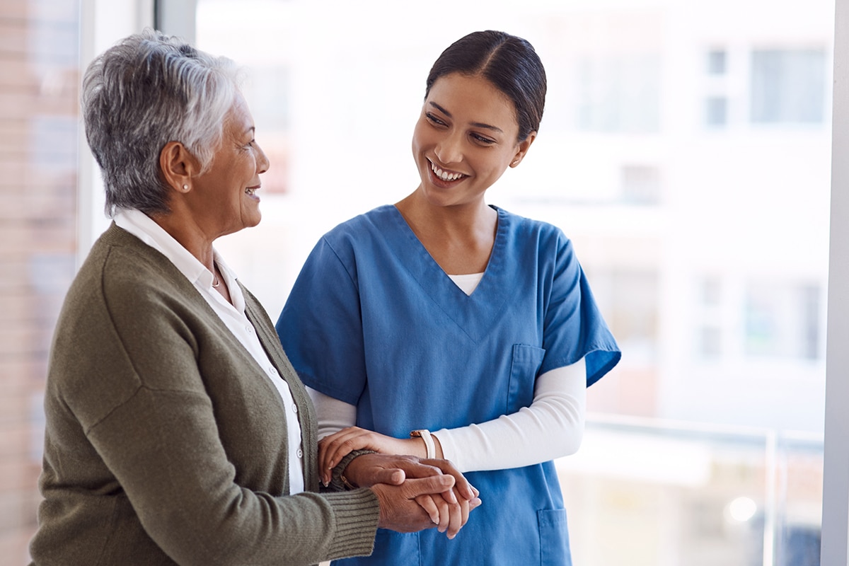 Support, caregiver with senior woman and holding hands for care indoors. Retirement, consulting and professional female nurse with elderly person smiling together for healthcare at nursing home.